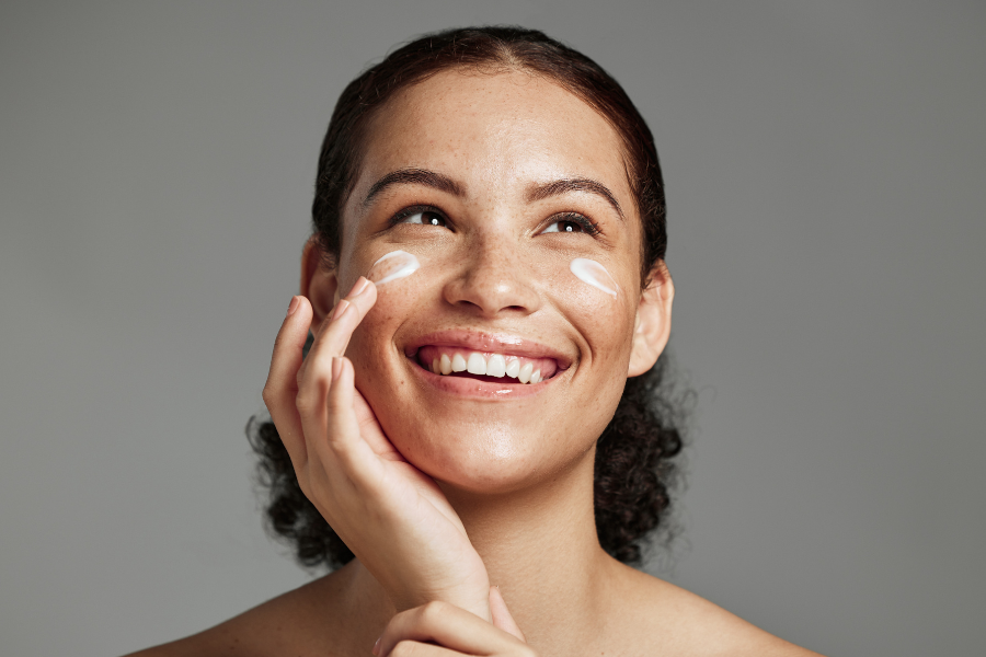 A woman smiles brightly as she applies cream to her cheeks, enjoying her skincare routine.
